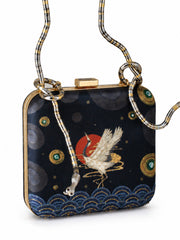 Close-up angled view of the Midnight Crane Clutch showing the embroidered crane, red sun, and gold circular motifs above the wave-patterned border.