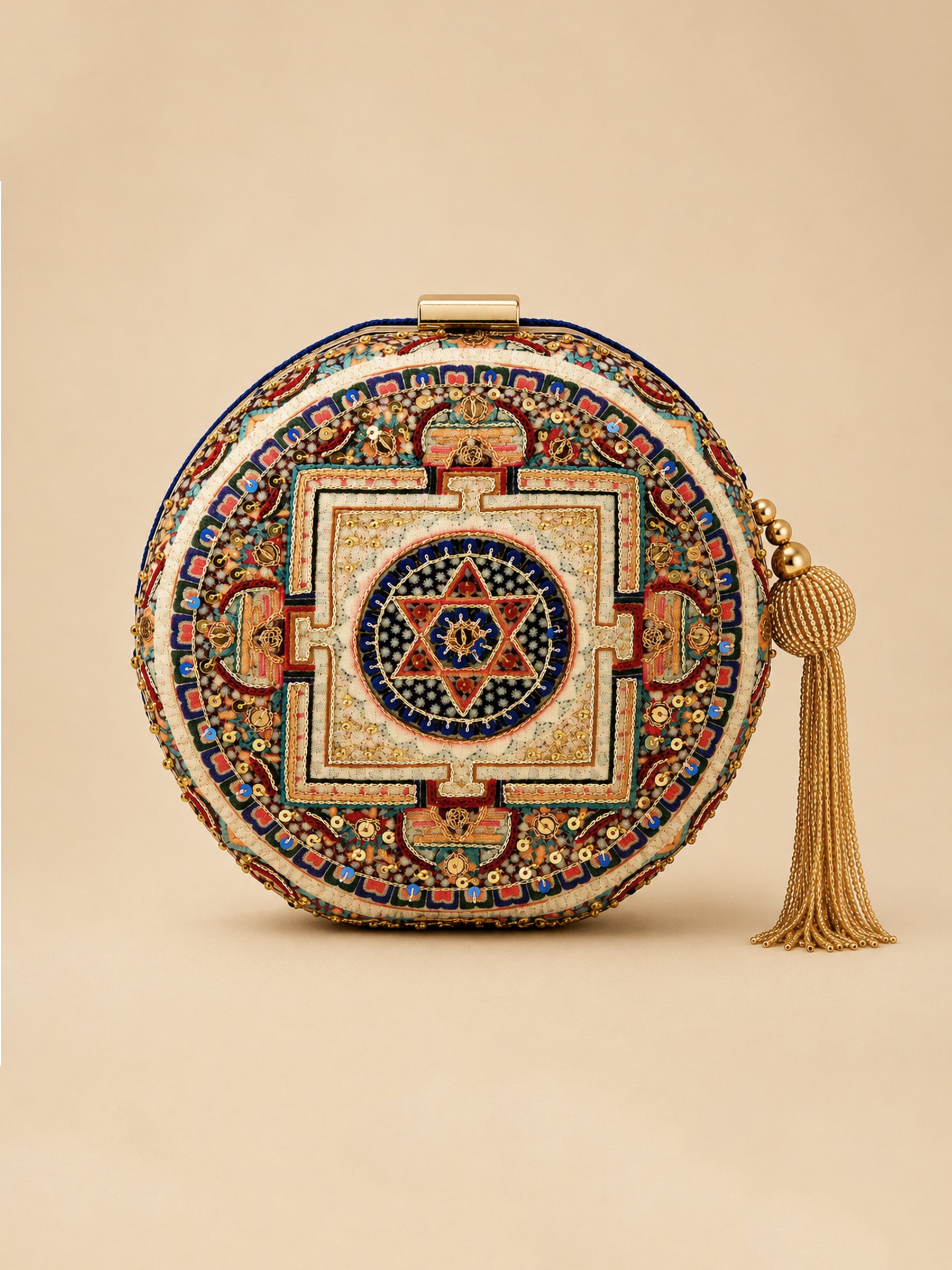 A round clutch bag with an intricate, colorful geometric and floral mandala design, featuring a gold-tone clasp and a matching gold tassel on a solid beige background.