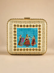 Saheliya Embroidered Clutch, a square box clutch in cream and turquoise velvet with a hand-embroidered panel depicting three traditional Rajasthani women in colorful attire, framed by a gold floral medallion border and a gold clasp.